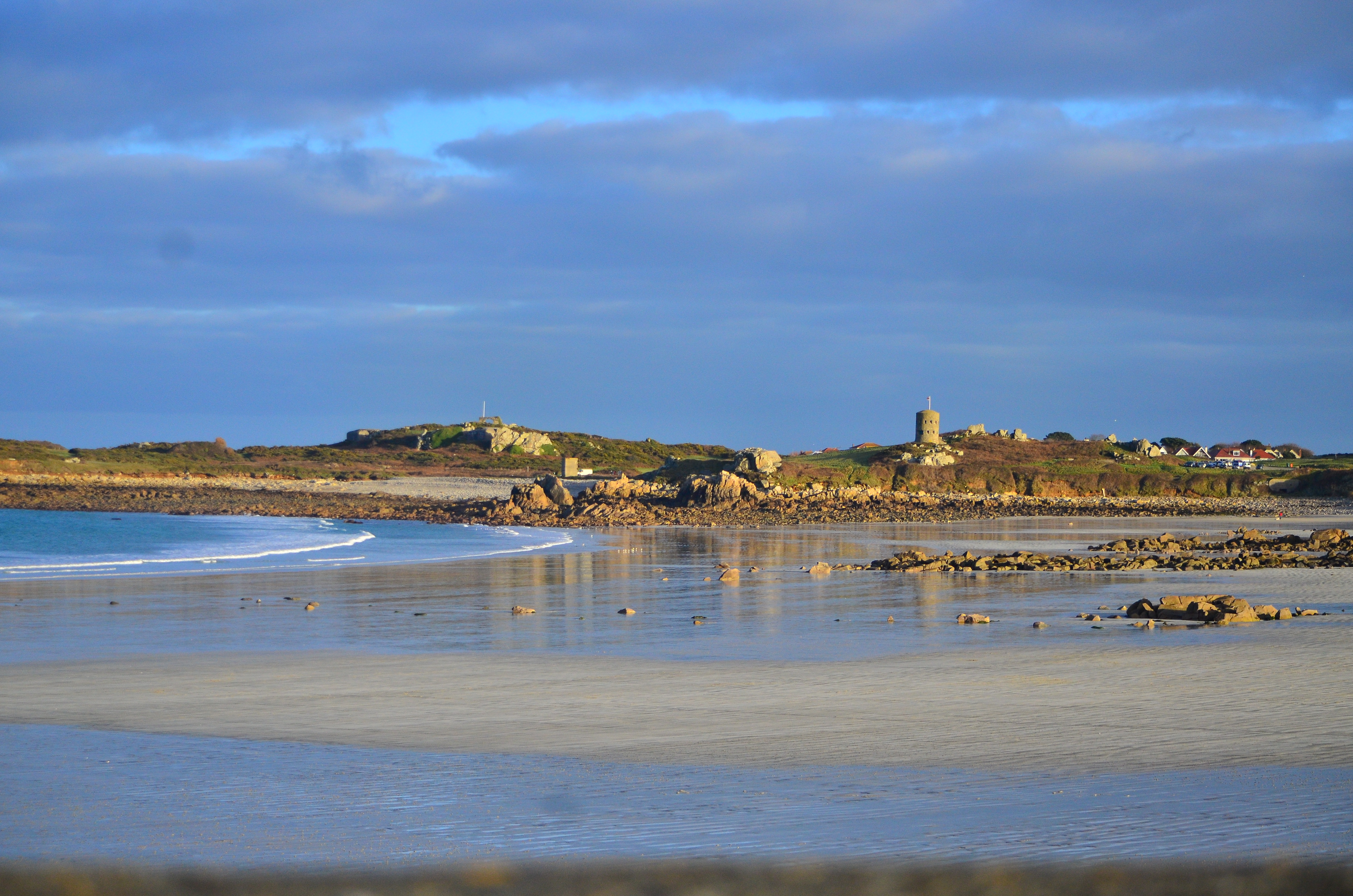The beach at low tide with gentle waves, wet sand, scattered rocks, and stone towers and coastal structures on the distant shoreline.